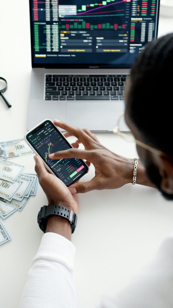 pexels-photo-6801872-6801872 A person trading stocks using a smartphone, showcasing financial data on mobile and laptop screens.